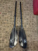 Full view of two black kayak or canoe paddle halves laid side by side on carpet, showing shafts and blades with OceanBroad logo.