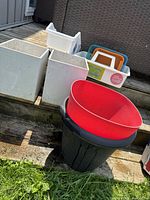 Outdoor photo showing two large white rectangular planters, a white plastic storage bin, multiple stacked colorful plastic trays, and a black planter with a red rim.