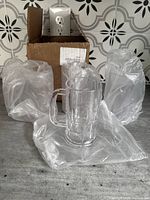 Four clear thick plastic drinking cups, all individually wrapped in plastic, arranged on a surface with a box in the background