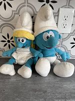 Photo of Smurfette and Smurf plush toys placed side by side showing overall size and condition on countertop against tiled wall.