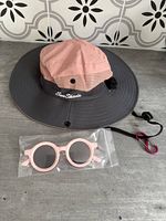 Toddler sunshade hat with wide grey brim, pink crown, mesh side panels, adjustable strap and red carabiner clip, light pink toddler sunglasses in front in bag.