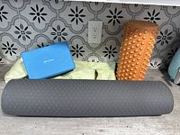 Yoga items including dark gray yoga mat rolled up, light green carry case, blue Renew yoga block, orange textured block, and dark gray foam roller on countertop.