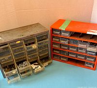 Two parts organizer cabinets, one metal and one orange plastic, with some drawers removed or open showing small watch and clock parts inside