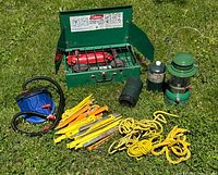 Photo displaying green Coleman Easi-Lite two-burner camp stove with red fuel tank in metal case, green lantern with metal handle, blue air pump with hose, yellow tent pegs, yellow ropes, and two propane cylinders arranged on grass.