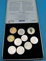 Overhead view of commemorative tin box containing 10 assorted Canadian coins arranged in two rows, with explanation text on the lid.