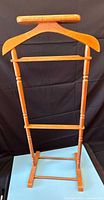 Front view of the wood valet stand showing its hanger top, multiple horizontal bars, and base with turned legs against a dark background.