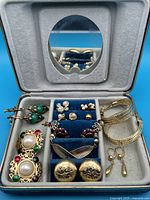 Earrings collection including large clip-on, green drop earrings, small studs, and gold-tone engraved earrings in a display case with mirror.