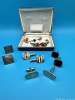 Photo showing all jewelry items including several pairs of cufflinks and some collar pins inside and outside the box, arranged on blue background.