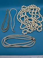 Four necklaces laid flat on blue background including a piled long strand, baroque pearl necklace, triple-strand pearls, and double-strand pearls