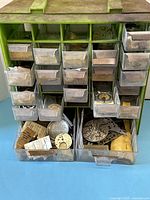 Front view of the green plastic organizer with multiple small drawers filled with assorted watch parts including movements, dials, and cases.