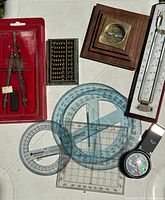 Wide shot showing all items: compass in wood base, thermometer in wood frame, abacus, protractor set, compass divider in red case, and step counter.