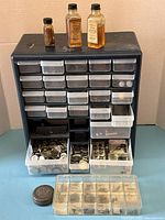 Full view of black metal organizer with 19 drawers and bottles of watch oil on top, various small watch parts in some drawers and containers at base.