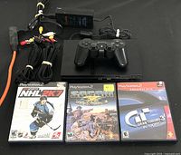 Front view of PlayStation 2 console with power adapter, AV cables, wired controller, and three video game cases laid out in front.