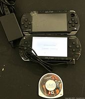 Two Sony PSP handheld consoles shown, one powered on showing initial setup screen, the other powered off, with power adapter and a UMD game disc on black surface.