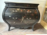 Front view of black lacquered chest of drawers showing curved bowfront design, three large drawers and two smaller drawers with painted bird, butterfly, and floral motifs and silver decorative handles.