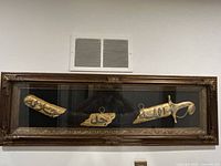 Full view of the framed wall hanging containing the sword and two scabbard pieces against a black background.