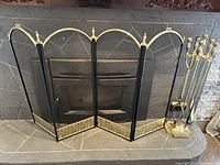 Foldable fireplace screen with four black mesh panels accented with gold arches and decorative trim at the bottom, set in front of a stone fireplace.
