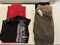 Five pairs of ladies dress pants in various colors including red, beige, black with patterns and olive, folded neatly showing back pockets and belt loops. Some pants show e-tags attached.