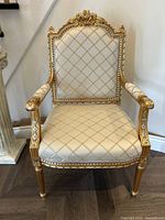 Front view of gold armchair showing detailed gold carved wood frame and cream diamond-tufted upholstery on seat and backrest.