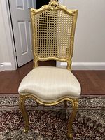 Full front view showing the gold-painted carved wooden frame, cane backrest, and upholstered beige seat.