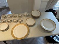 Full view of multiple pieces of white china with gold accent including coffee cups, plates, bowls, and serving platters on a table.