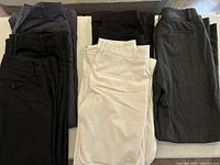Seven pairs of ladies dress pants arranged in stacks on a floor showing multiple colors including black, gray, navy, and white.