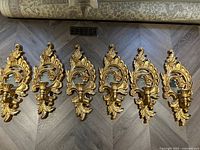 Six ornate gold leaf candle wall sconces laid out on floor, visible decorative detailing and curved candle holders.