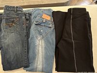 Three pairs of pants displayed side by side: dark denim jeans with embroidered pocket, light blue jeans with buttoned detail, and black leggings with white seam.