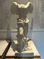 Front view of white winged angel statue with draped garment and no head or arms, shown on a base with ornate furniture and wallpaper background.