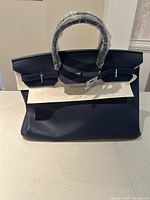 Front view of blue leather purse, showing flap closure with silver turn-lock and handles wrapped in protective plastic.