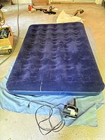 Photo showing the dark blue air mattress fully inflated on a blue tarp with the electric air pump and power cord in front of it. Background shows equipment and garage floor.