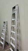 Side view of two aluminum ladders leaning against a wall, showing the height and step spacing of the main ladder.