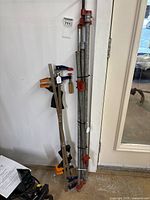 Photo showing several metal bar clamps including large pipe clamps with red iron clamping sections leaning against a white wall near a door.