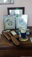 Three wooden bathroom brushes with a round mirror, two framed blue floral art prints, and some decorative items arranged on a wooden table.