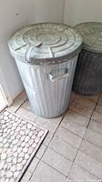 Photo of large galvanized metal trash can with lid and handle, placed on a tiled floor by a door mat.