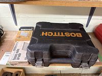 Closed black Bostitch branded hard plastic carry case with some boxes of finishing nails placed beside it on a shelf.