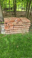 Stacked red clay bricks in wooded area with grass in front. Bricks have some wear and chipping.