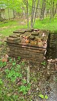 Photo of a large stack of red clay bricks and some clay tiles on top, placed outdoors on dirt and surrounded by greenery and trees.