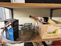 Shelf with wooden crate filled with car cleaning brushes, spray bottles, and other cleaning supplies along with a boxed 4 ton hydraulic jack and metal hand winch.
