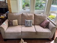 Front-facing view of the entire sofa with three decorative accent pillows and three back cushions on tiled floor in front of windows.