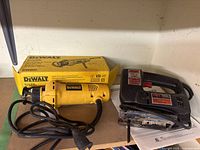 DeWalt cut-out tool DW660 in front of its original yellow box alongside black Skil jig saw on a shelf.