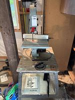 Front view of grey Delta band saw on stand with blade guard and fence, manual visible on lower shelf