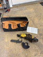 Yellow DeWalt DW818 angle grinder with power cord coiled, side handle detached, safety guard and user manual next to it, all sitting on a workshop floor. Black and orange carry bag positioned behind the tools.