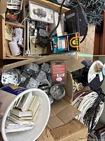 Two cardboard boxes with mixed electrical hardware including metal electrical boxes, switches, and unused packaged items, some cords coiled nearby