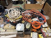 Overview photo showing a variety of coiled extension cords in white, black and orange, several power bars, a portable work light with guard, outlet adapters, and other electrical items on a table.