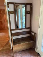 Front view of wooden entryway bench showing seat with storage box design and upper section with six brass coat hooks and central mirror.