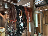 Wide view of multiple extension cords hanging on wooden beam in a shed or garage setting, showing various colors and coiling.