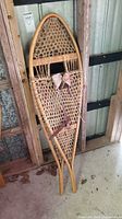 Pair of vintage wooden snowshoes leaning against wooden paneling, showing full frames, rawhide woven webbing and leather bindings with some wear visible.