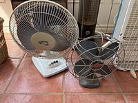 Set of two fans photographed on tiled floor, highlighting both the vintage Electrohome metal fan and the white modern plastic fan with buttons.