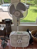 Wide shot of three fans placed near a window including the pedestal fan at the back, the box fan in front, and a small vintage fan to the left.